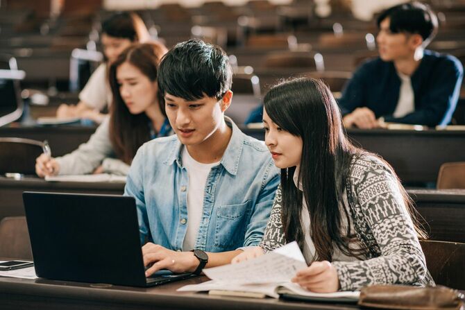 大学の教室内で男女の学生が同じノートパソコンを見ている