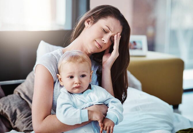 赤ちゃんを抱きながら、こめかみと額を押さえて困った表情を浮かべる母親