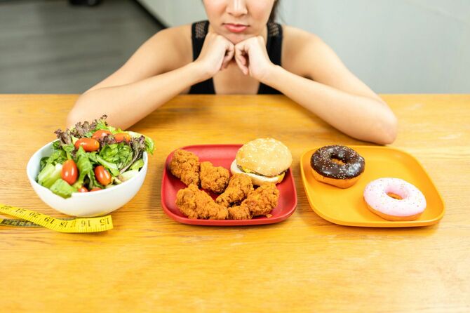 サラダボウル、ジャンクフード、ドーナツの乗った皿を見て悩む女性