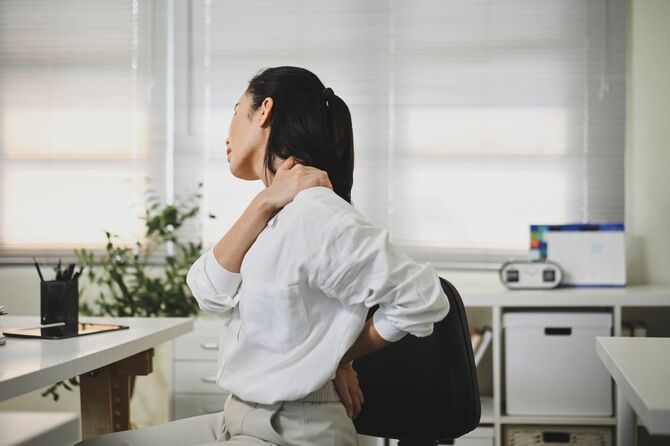オフィスの自席で肩の張りを感じて、手を当てている女性