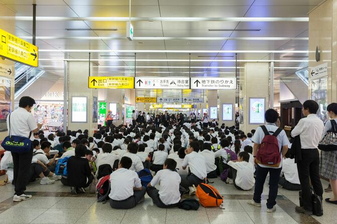 東京駅の通路いっぱいに並んで座り、先生の話を聞いている修学旅行中の生徒たち