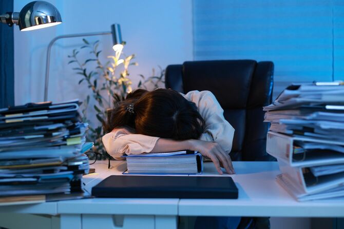 デスクライトはついたまま、書類の山の間で机に突っ伏して仮眠をとっている疲れた女性