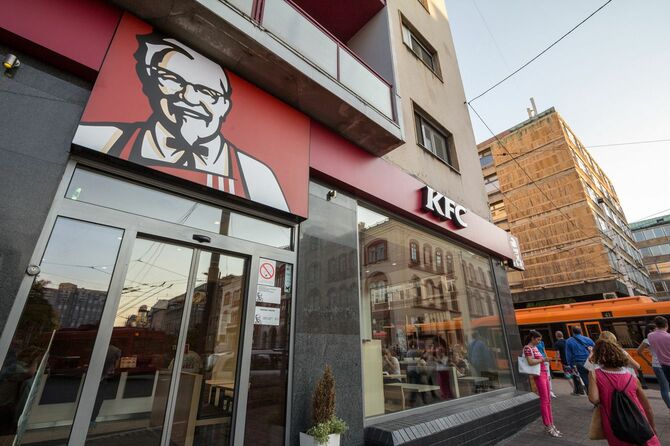 セルビアのベオグラードの街角にある、カーネル・サンダースがシンボルのKFCの看板の前を行き交う人々。KFCは、フライドチキンを専門とするアメリカのファストフードレストランチェーンで、世界第2位のレストランチェーン