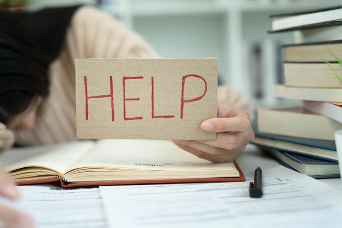 「HELP」の字が書かれた段ボール紙を掲げながら、机に突っ伏している女性