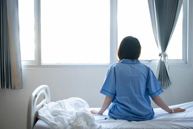 病院のベッドに座り、外を眺める女性の後ろ姿